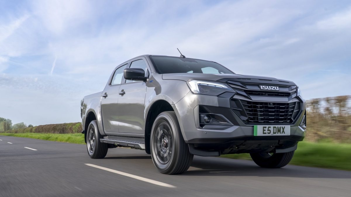 Gray Isuzu pickup truck in motion on a two-lane road under a blue sky, from a low-angle side view.