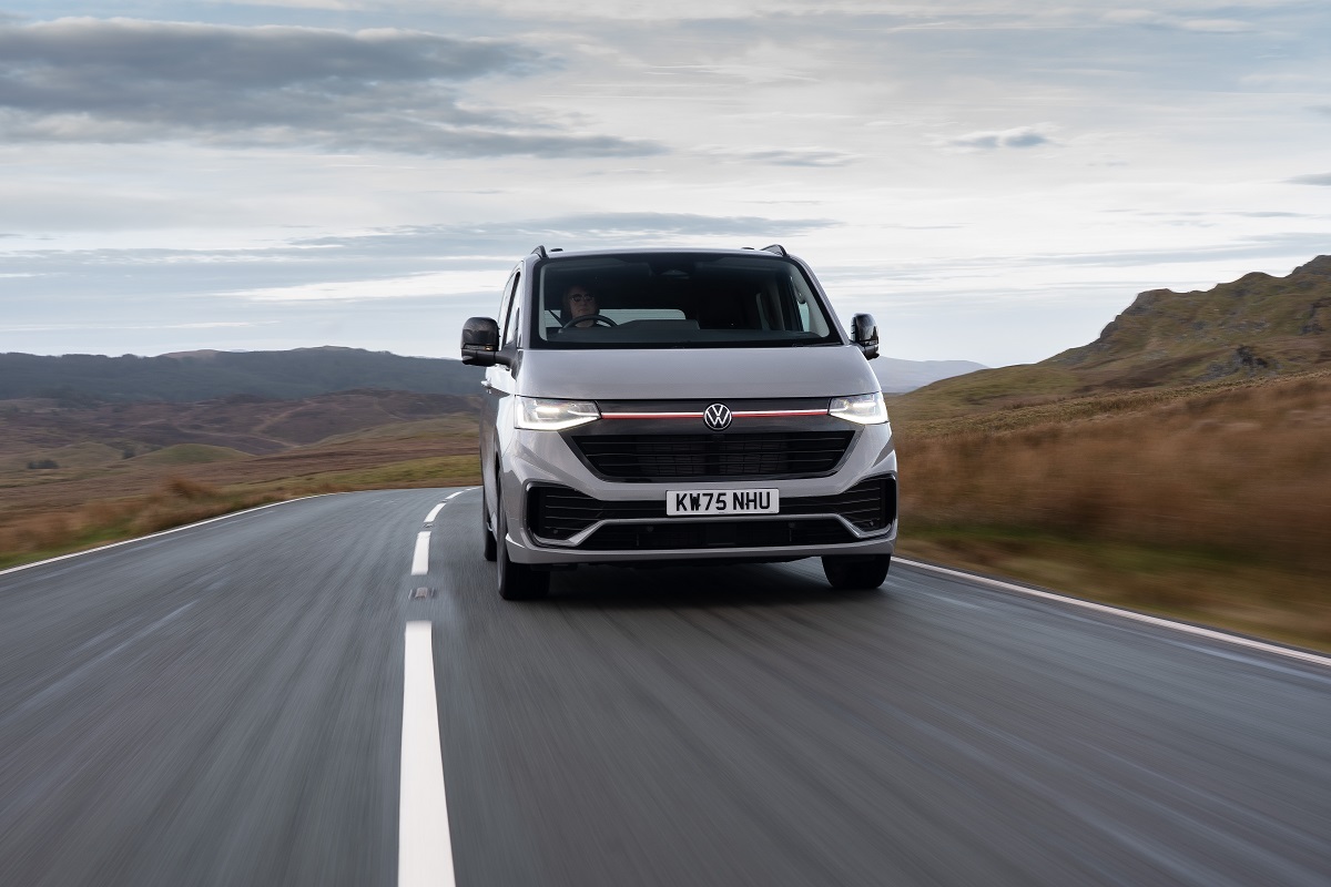 Silver Volkswagen van driving toward the camera on a rural two-lane road with open fields and hills in the distance.