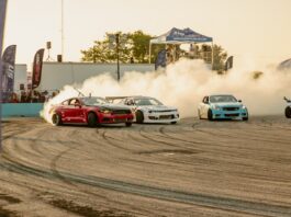 Red drifting car leads a pack on a smoky racetrack, white and blue cars following as spectators watch from behind barriers under a pavilion.