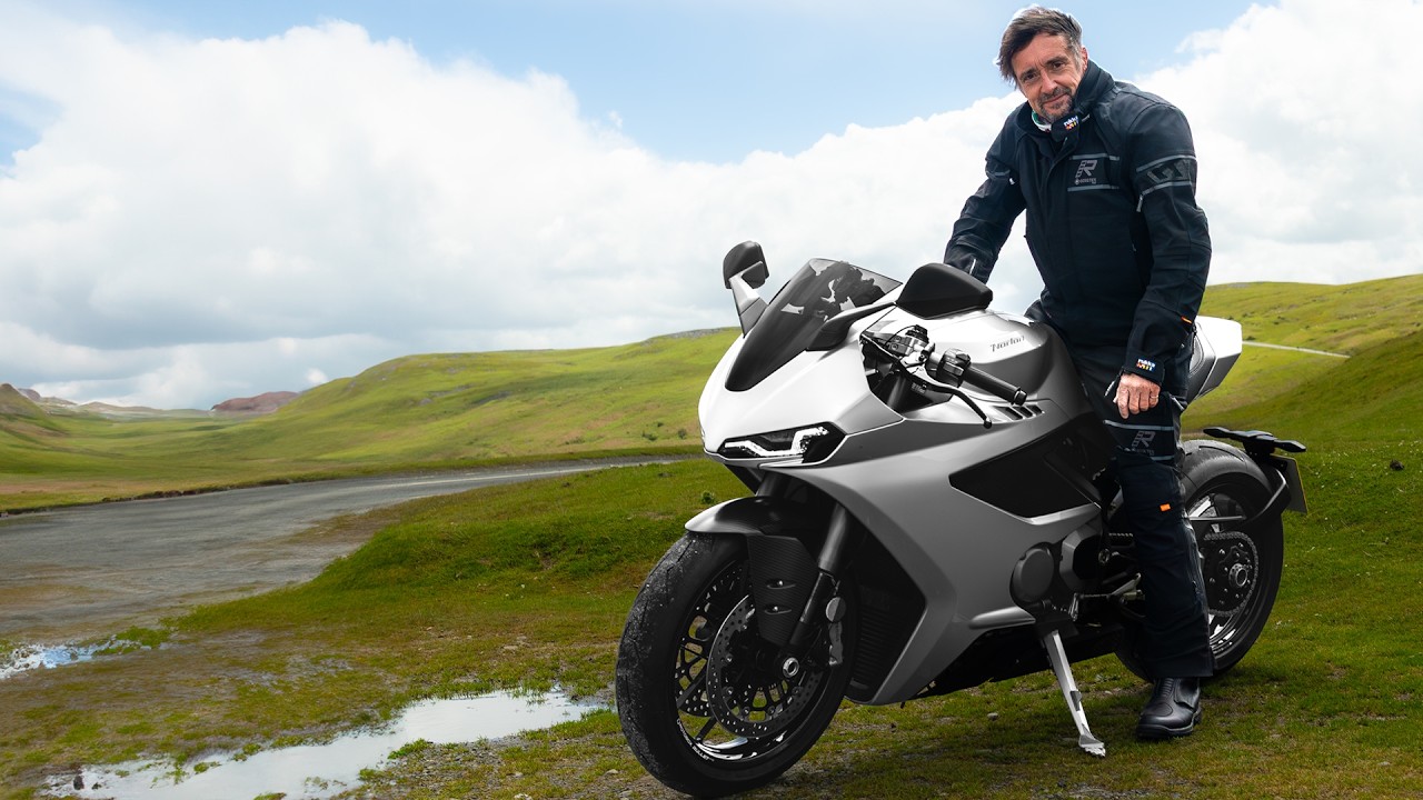 Man in riding gear standing beside a silver sport motorcycle on a grassy hill road.