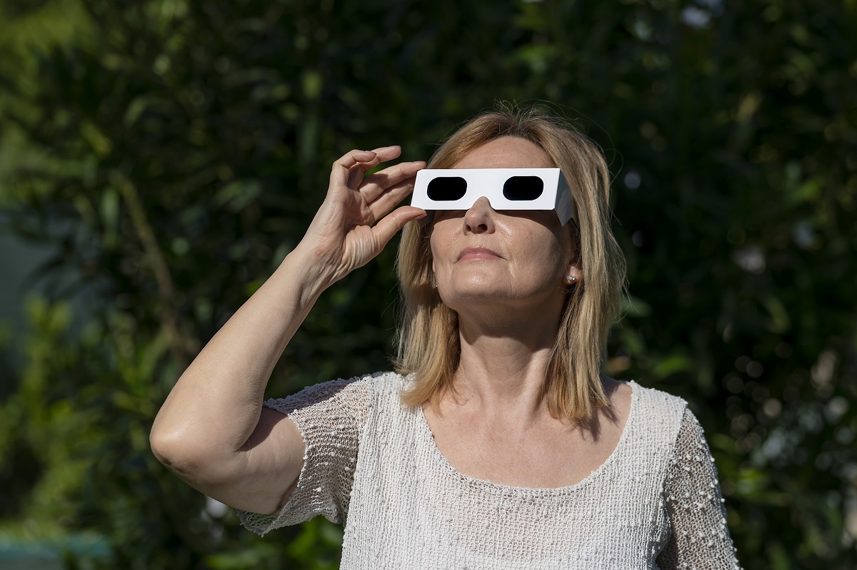 Older woman outdoors wearing white cardboard 3D glasses, looking upward in a garden, sunlight on her face.